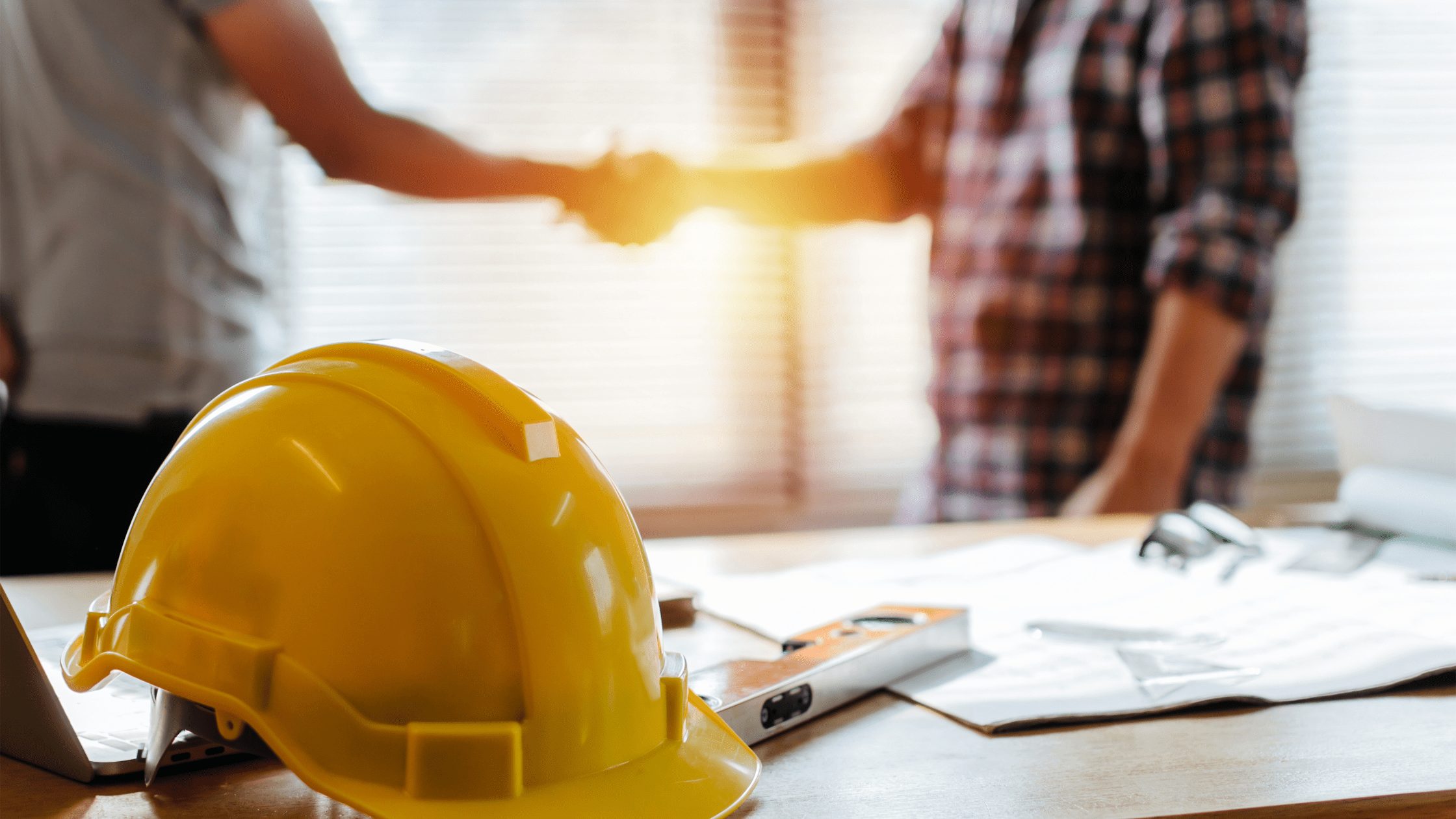 Yellow hard hat and building plans on a table, with two people shaking hands in the background.