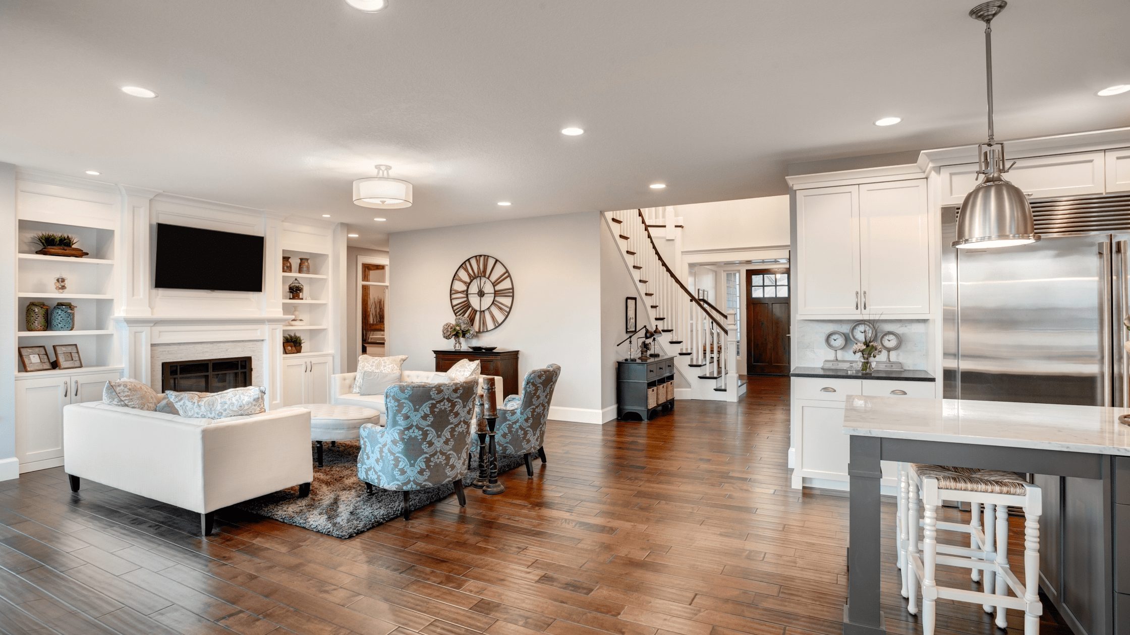 Open-plan living room and white kitchen with a fireplace, built-in shelves, blue patterned chairs and wood flooring.