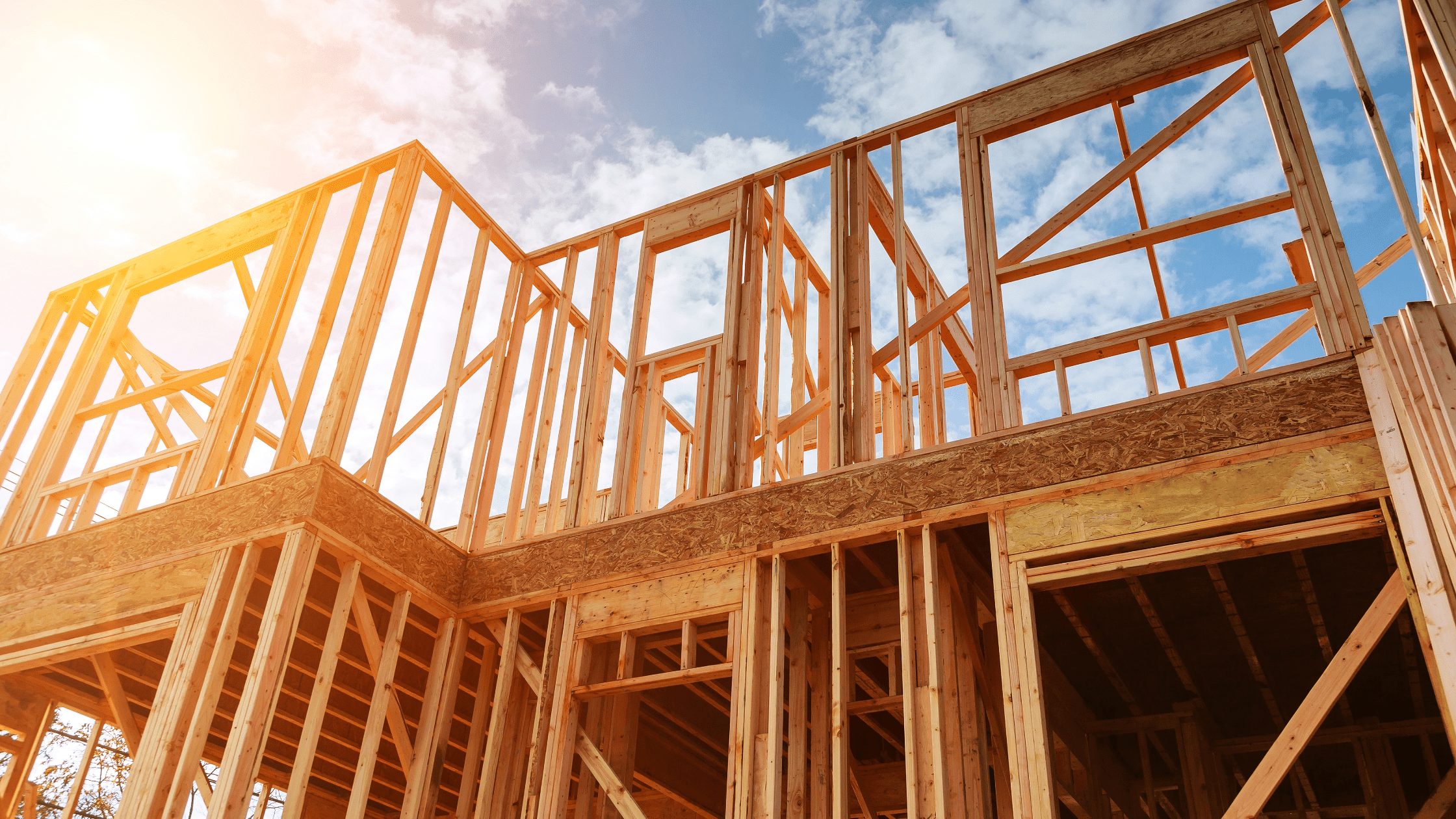 Exposed wood framing of a two-story house under construction, with open window frames against a blue sky.