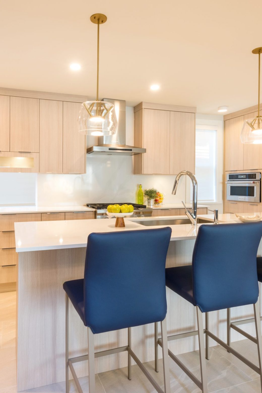 Kitchen with pale wood cabinets, a white island, blue counter stools and stainless steel appliances.