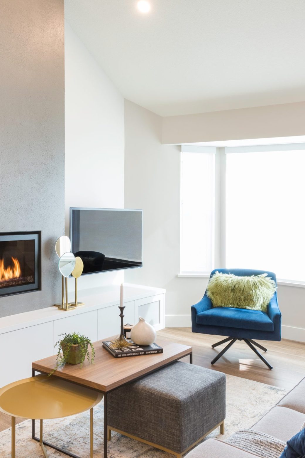 Living room with a wide fireplace, floating white cabinets, gray sofa and blue chair beside a bay window.