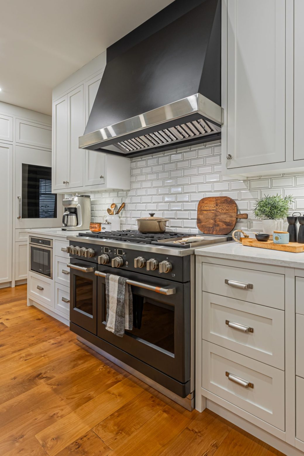 Dark kitchen range and matching hood framed by white cabinets and a white tile backsplash above wood flooring.