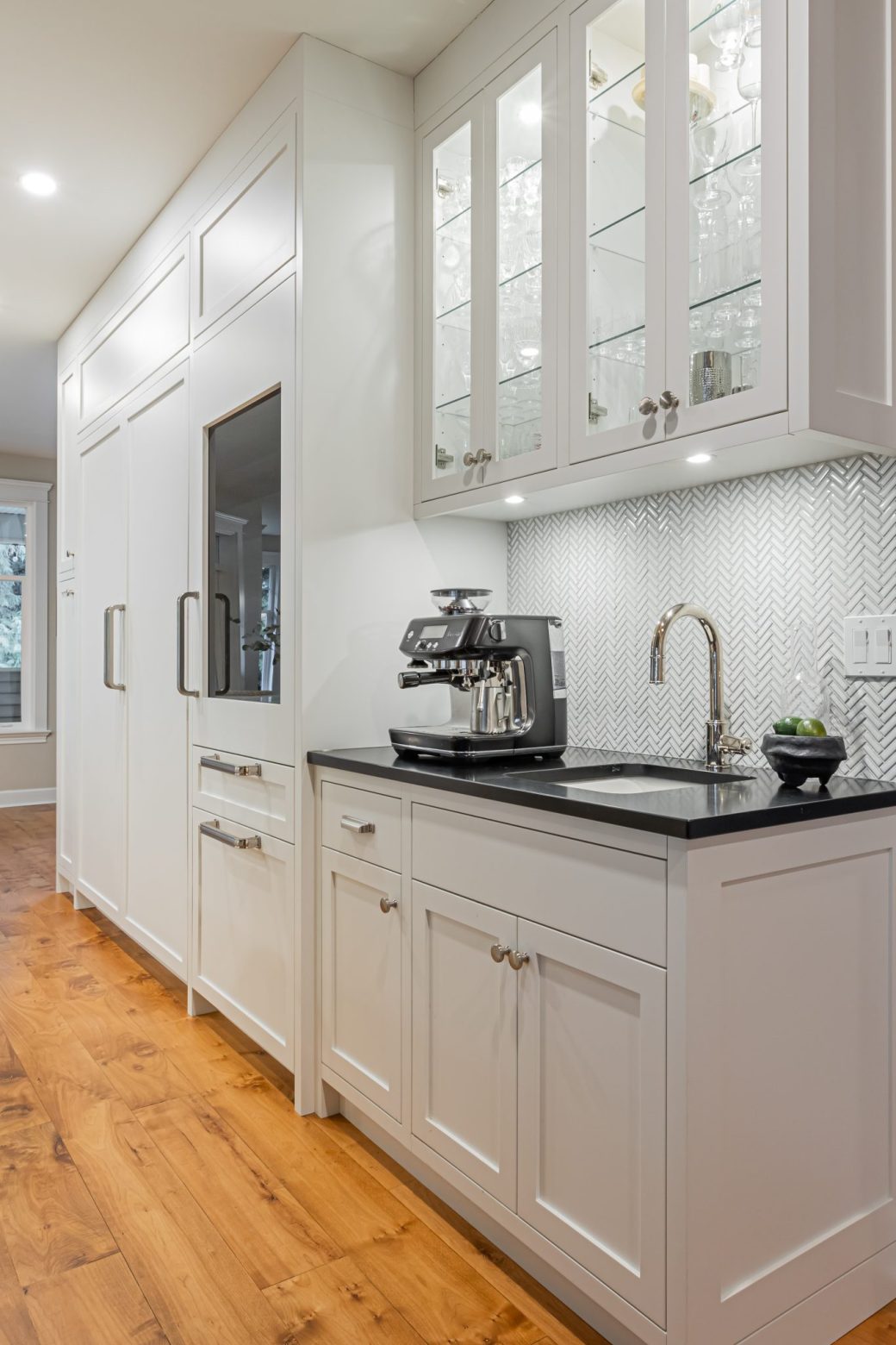 White built-in coffee station with a dark counter, sink, herringbone backsplash and illuminated glass-front cabinets.