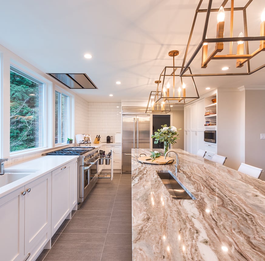 White kitchen with a veined island, trough sink, glass lantern pendants and a wide window.