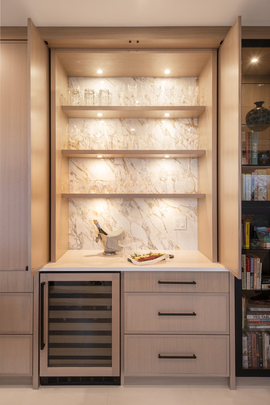 Open cabinet doors reveal illuminated glassware shelves, a veined backsplash, drawers and a built-in wine fridge.