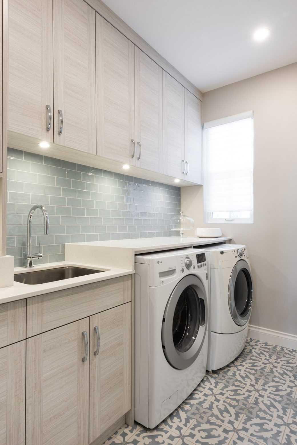 Laundry room with pale cabinets, a sink, front-loading appliances and a patterned tile floor.