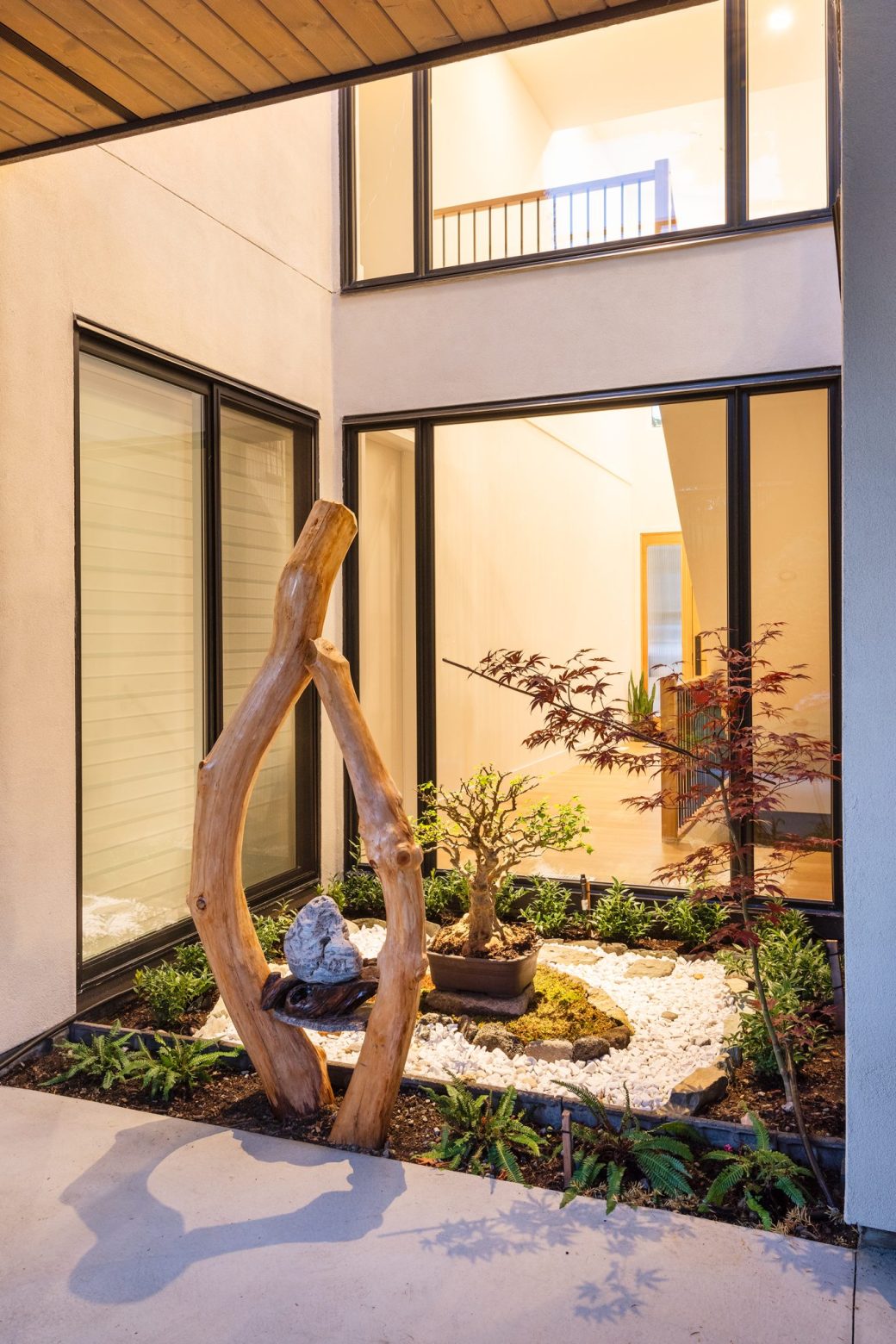 Small courtyard garden with sculptural wood, gravel and plants framed by tall windows.