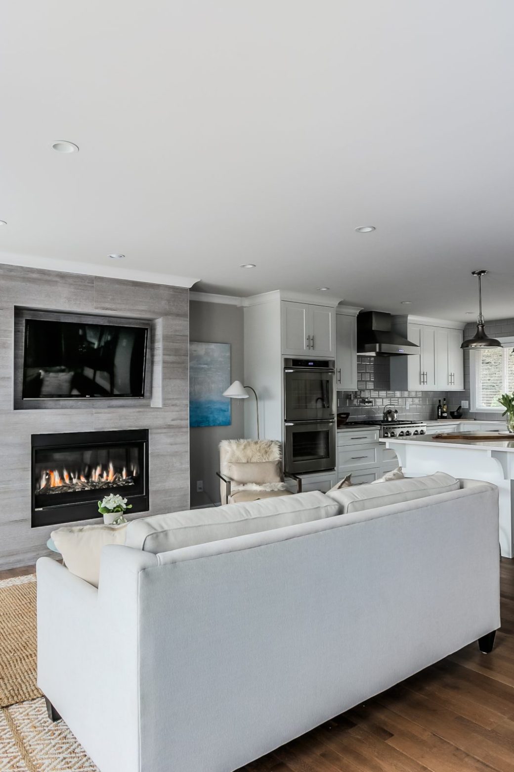 Open living room and white kitchen with a tiled fireplace, wall-mounted television and window bench.