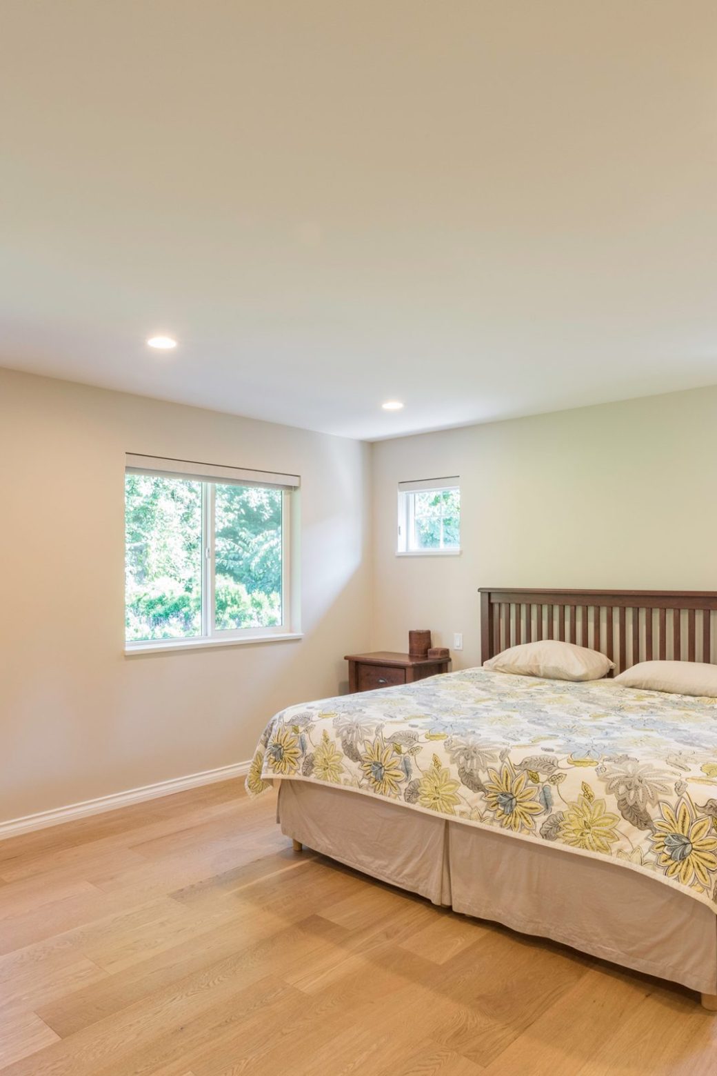 Bright bedroom with a wood bed frame, floral bedding, multiple windows and light wood flooring.