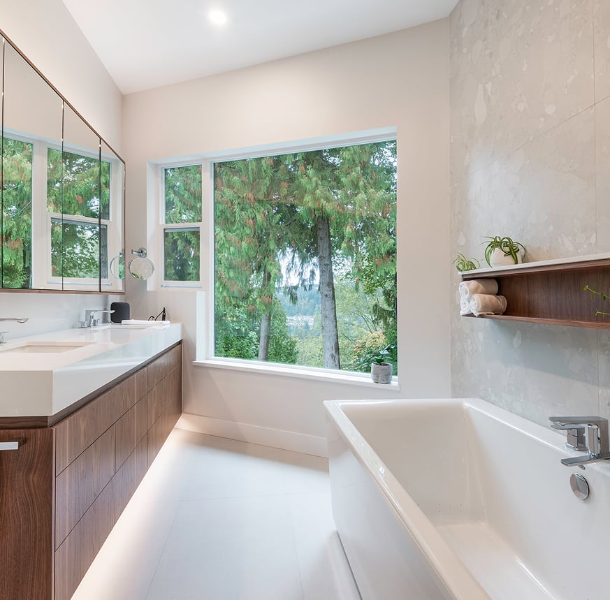 Wood bathroom vanity and mirrored cabinets beside a freestanding white tub with a leafy window view.
