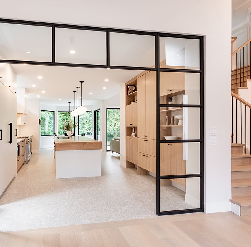 Kitchen with a white island and wood cabinets behind a black-framed glass partition beside a wood staircase.